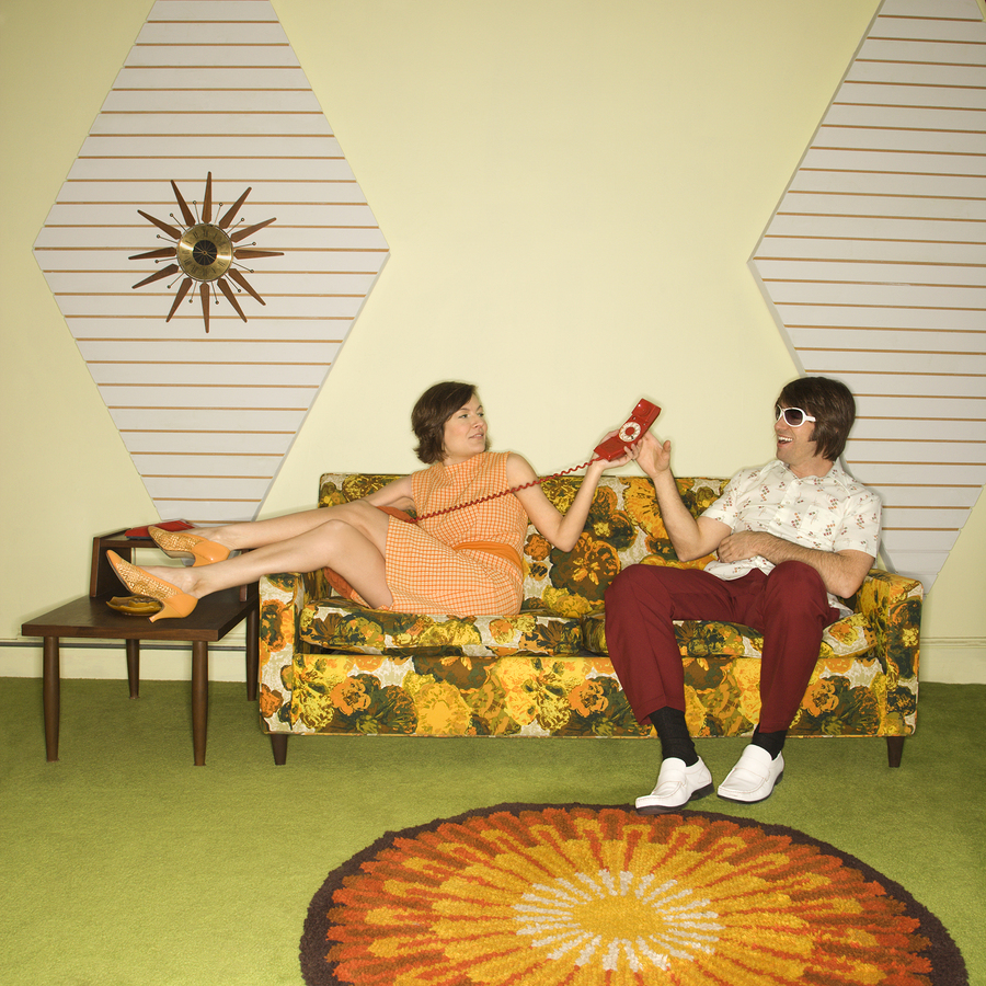 Caucasian mid-adult woman passing phone to Caucasian mid-adult man wearing sunglasses sitting on sofa.